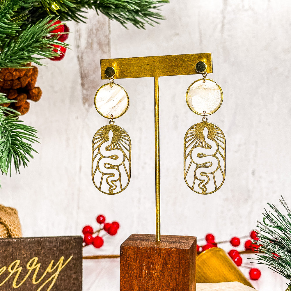 The Dappled Wood Ivory Coast framed circle earrings with a delicate snake sun charm, crafted from polymer clay with gold-tone details. A bold yet lightweight statement piece.