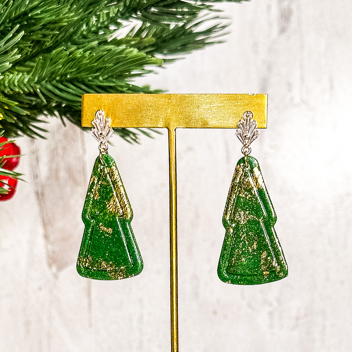 The Dappled Wood Noelle Christmas tree earrings, displayed on a gold stand, featuring hand-embossed golden details on a vibrant evergreen base. A bold and elegant holiday accessory.