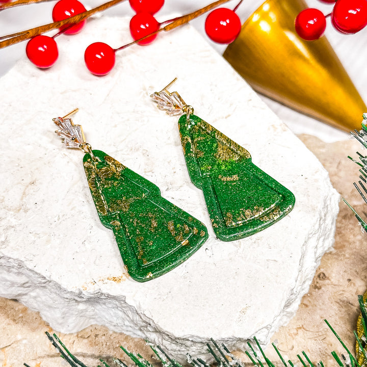 The Dappled Wood Noelle Christmas tree earrings, handcrafted from polymer clay in a deep green hue with intricate gold detailing. A festive statement piece for the holiday season.