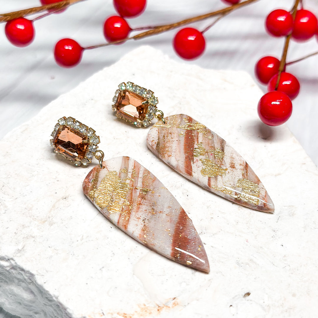 The Dappled Wood Patti earrings, inspired by peppermint bark, featuring marbled white and red polymer clay with delicate gold leaf accents. Finished with a sparkling champagne glass gem post for an elegant holiday statement.