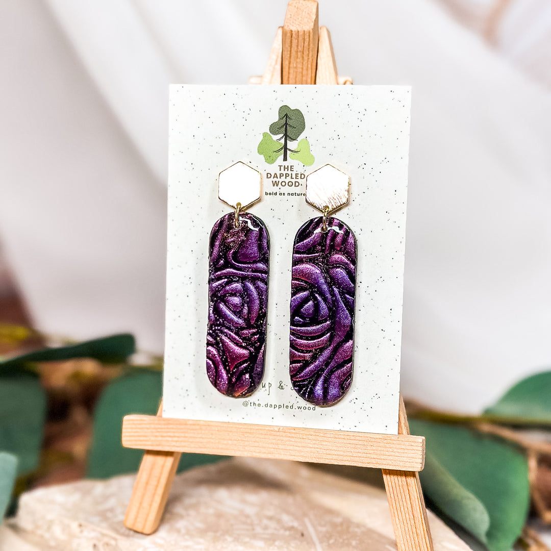 The Dappled Wood Carla rose-embossed dangle earrings, handcrafted in deep purple polymer clay with floral detailing. Suspended from brushed gold hexagon posts, these lightweight statement earrings add an elegant touch to any outfit.