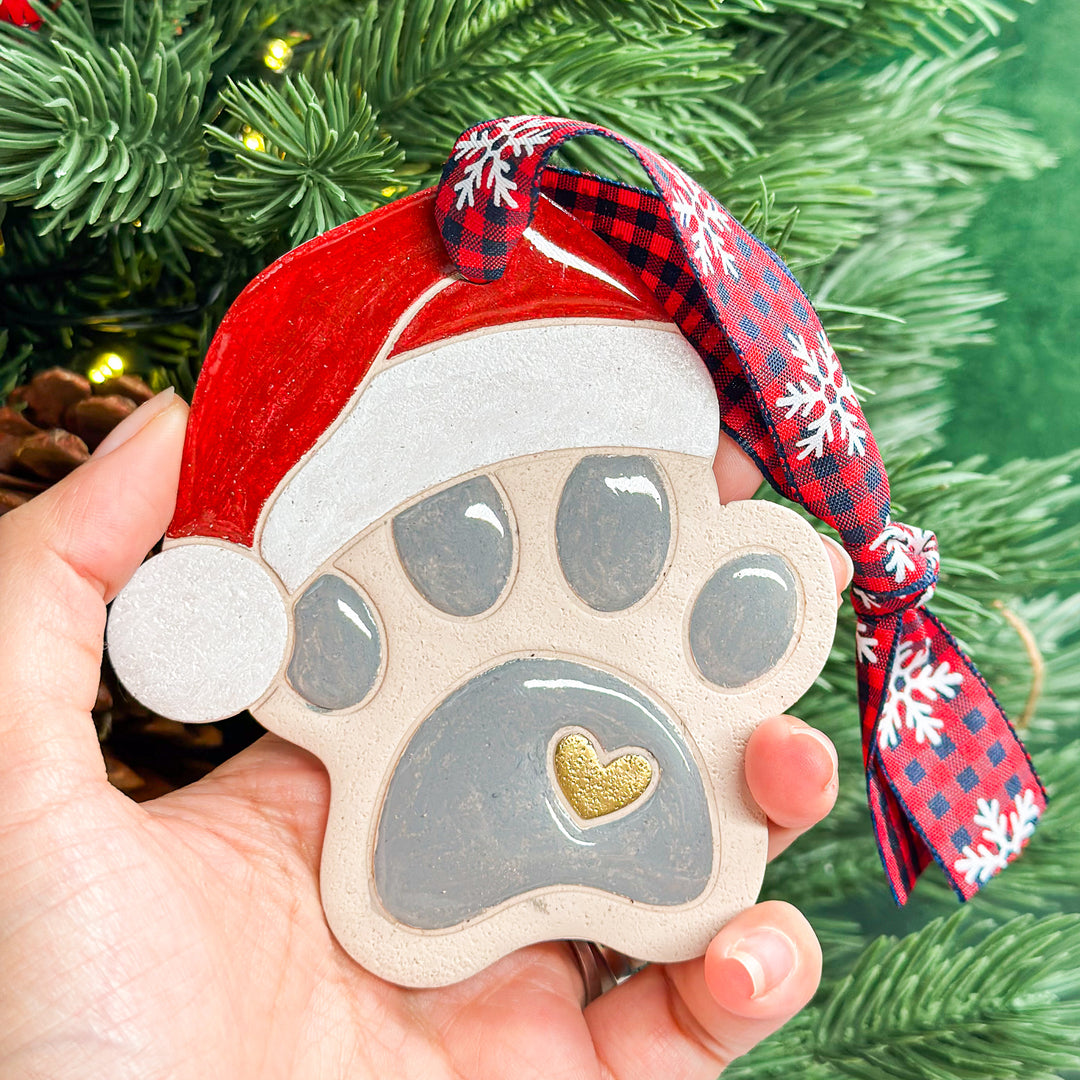 Hand holding a paw print ornament with a red Santa hat, a festive holiday keepsake. 2025.