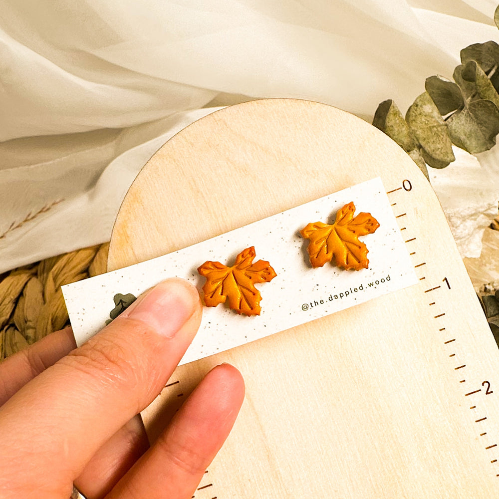 Orange Briar maple leaf stud earrings on an earring card beside an inch ruler