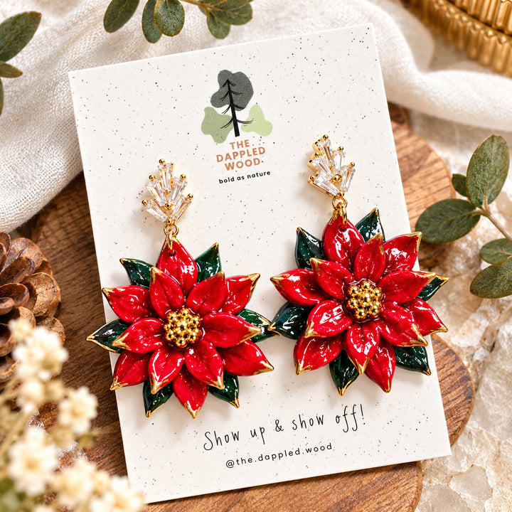 Carmela red sculpted poinsettia statement earrings with dark green leaves, gold bead centers and crystal fan posts on a white jewelry card