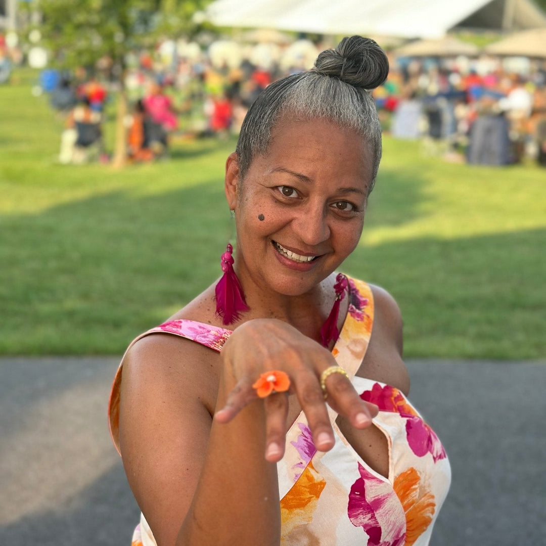 Customer smiling at an outdoor event while showing The Dappled Wood Isadora flower ring in orange, paired with a floral dress and tassel earrings.