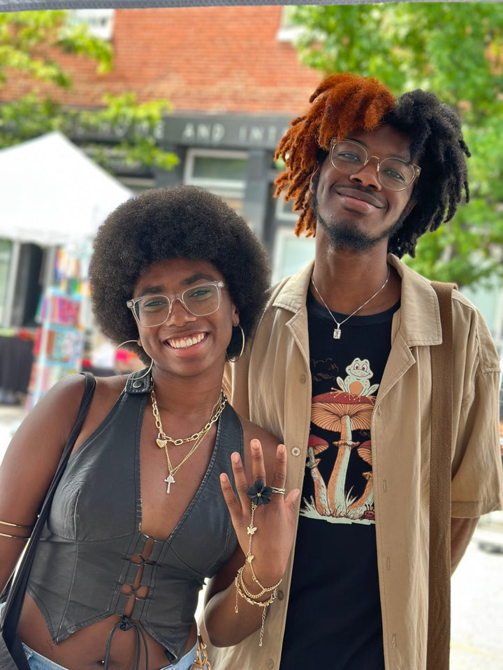 Two customers smiling at an outdoor market, one showcasing The Dappled Wood black Isadora flower ring while wearing a black outfit and layered jewelry.