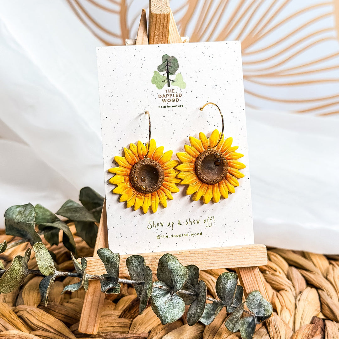 Clara handmade sunflower earrings by The Dappled Wood, hypoallergenic and lightweight, handmade in Baltimore displayed on branded card with eucalyptus greenery and wicker background