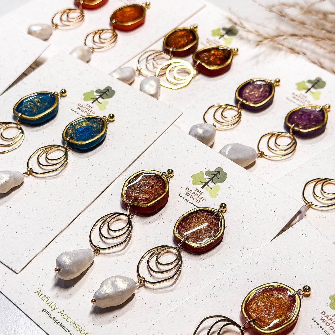 The Dappled Wood Fall 2025 collection of Rhea earrings in multiple color variants, featuring faux baroque pearls and statement drop designs displayed on branded earring cards.
