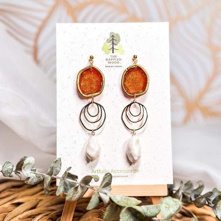 The Dappled Wood Fall 2025 Rhea earrings in pumpkin orange with faux baroque pearls, displayed on a branded earring card, styled as a statement accessory for fall outfits, autumn gatherings, and Thanksgiving looks.