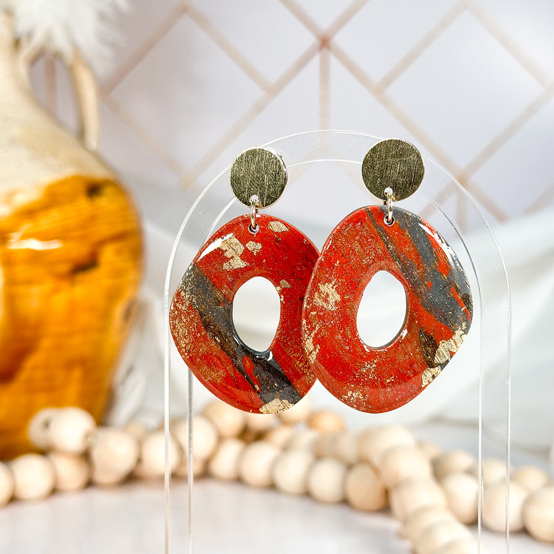 The Dappled Wood Brick Red and Gold statement earrings, handcrafted from faux stone polymer clay with a marbled red and black design, accented with gold leaf. Suspended from textured brass studs for a bold, artistic look.