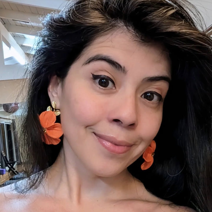Customer wearing The Dappled Wood Flora earrings in orange while smiling outdoors at a local market. The bold flower design highlights the lightweight statement jewelry style of the brand.