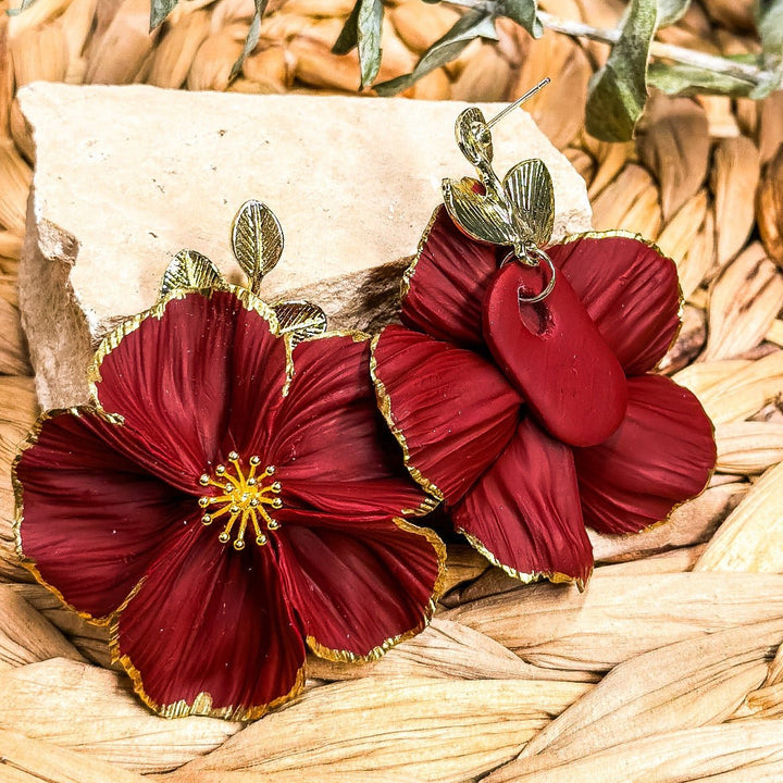 The Dappled Wood Flora earrings in deep red with gilded petal edges shown on a woven display surface.