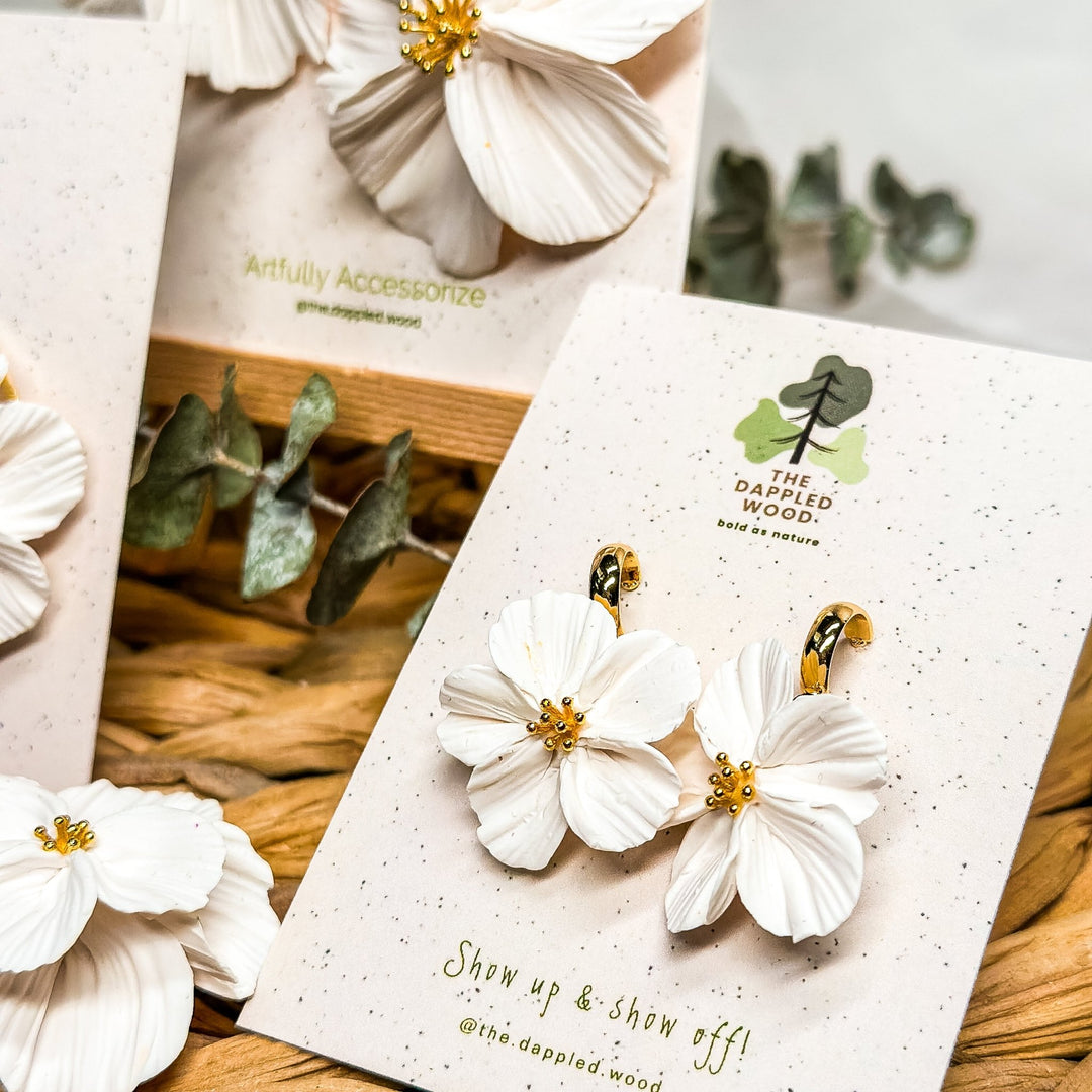 Closeup photo of the Florita mini flower earrings from The Dappled Wood. Small white floral earrings with gold centers displayed on a branded earring card with greenery in the background.