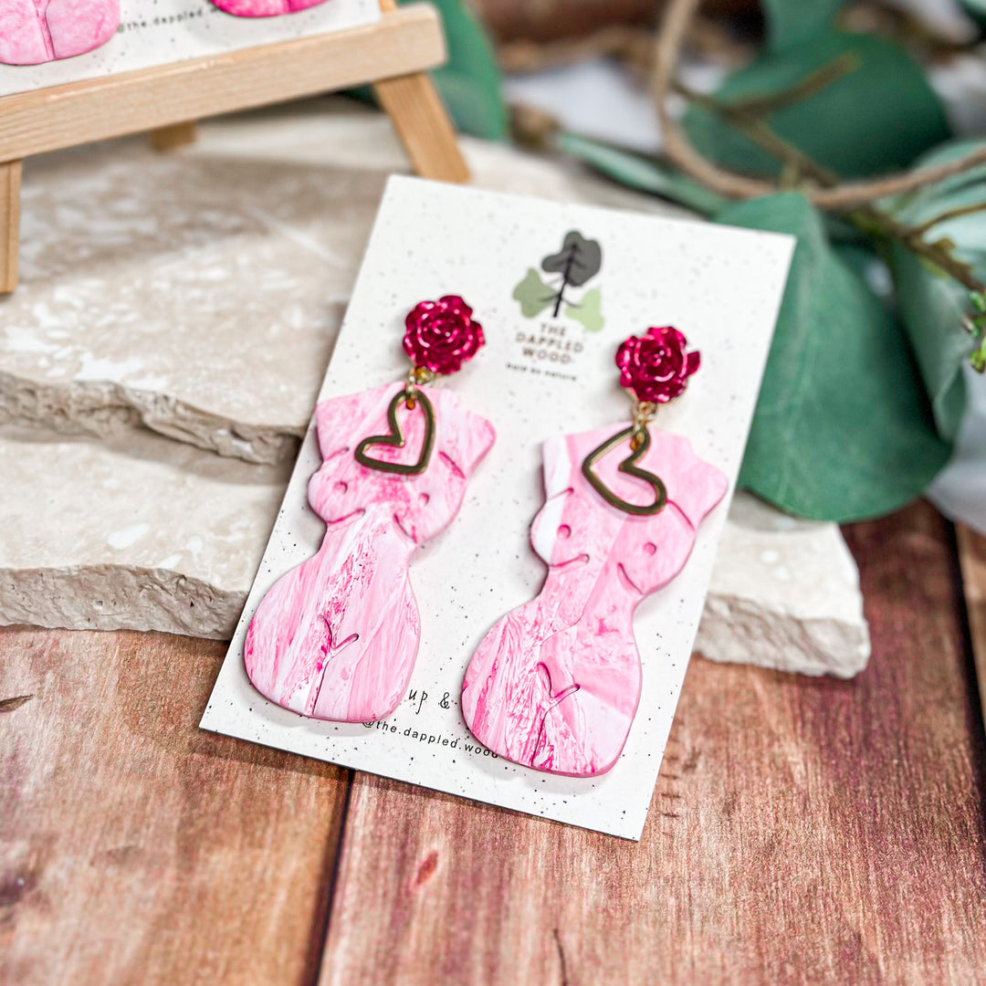 The Dappled Wood Frenchie earrings in Rosebud. Pink female figure statement earrings with deep red floral accents, handcrafted from polymer clay. Lightweight and hypoallergenic jewelry.