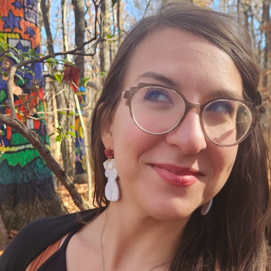 Customer wearing the Frenchie earrings by The Dappled Wood, featuring a white female figure silhouette with red accents while posing outdoors among colorful painted forest art.
