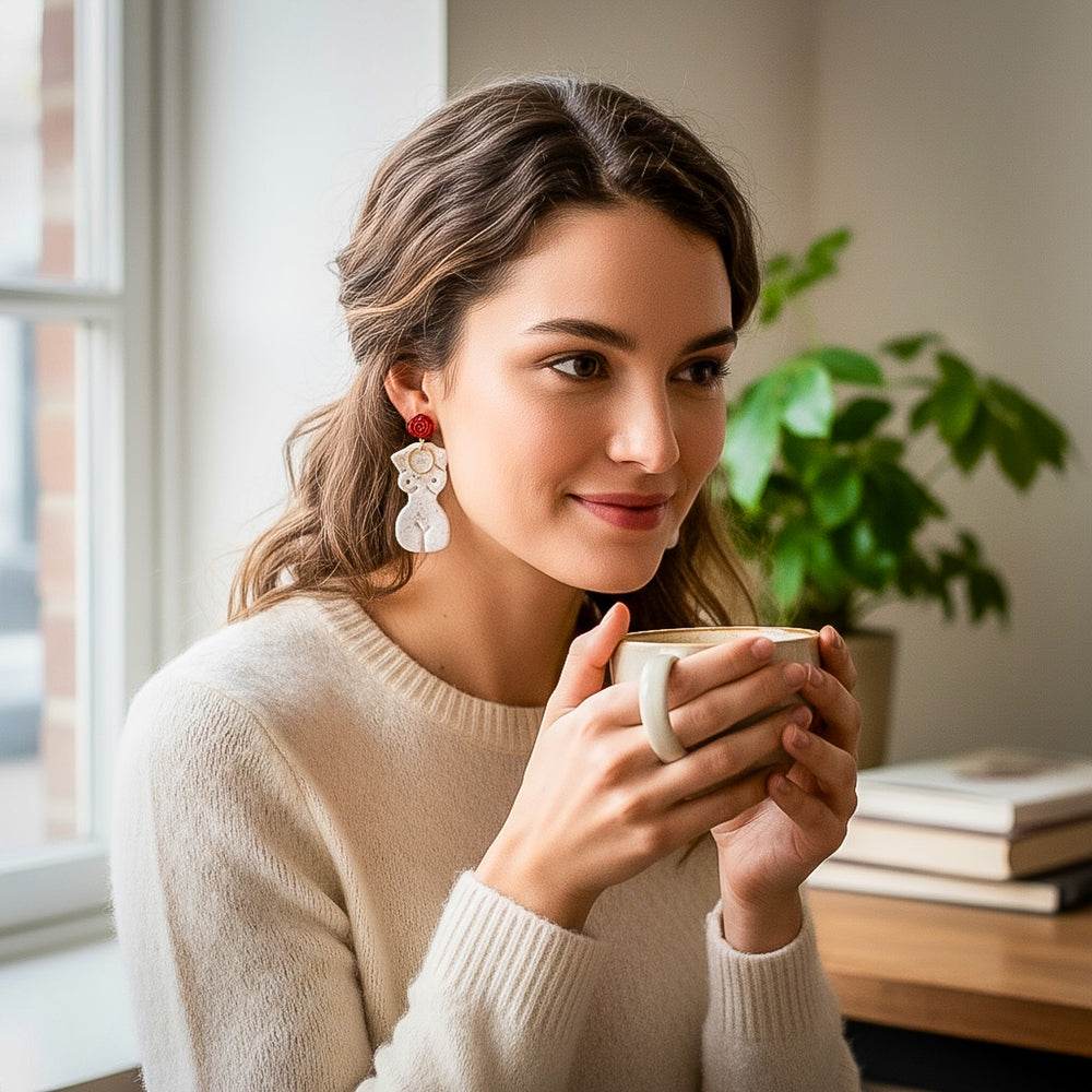 Model wearing the Frenchie earrings by The Dappled Wood, featuring a white female silhouette design with red accents while enjoying a warm drink in a cozy indoor setting.
