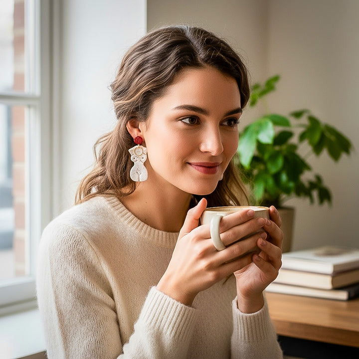 Model wearing the Frenchie earrings by The Dappled Wood, featuring a white female silhouette design with red accents while enjoying a warm drink in a cozy indoor setting.