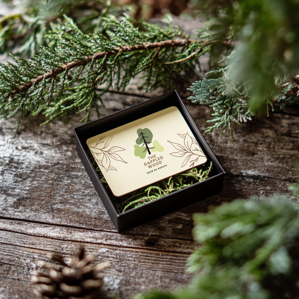 The Dappled Wood gift card displayed in a black box with natural moss, set on a rustic wooden surface surrounded by evergreen branches. A perfect gift for handmade jewelry lovers to choose their favorite nature-inspired accessories.