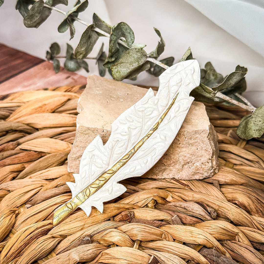 Gilded Feather Brooch by The Dappled Wood in sculpted white with gold detailing, shown in a styled close up on a natural textured background.