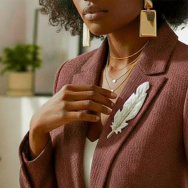 The Dappled Wood Gilded Feather Brooch in white and gold worn on a textured mauve blazer, shown in a polished lifestyle photo with layered gold jewelry.