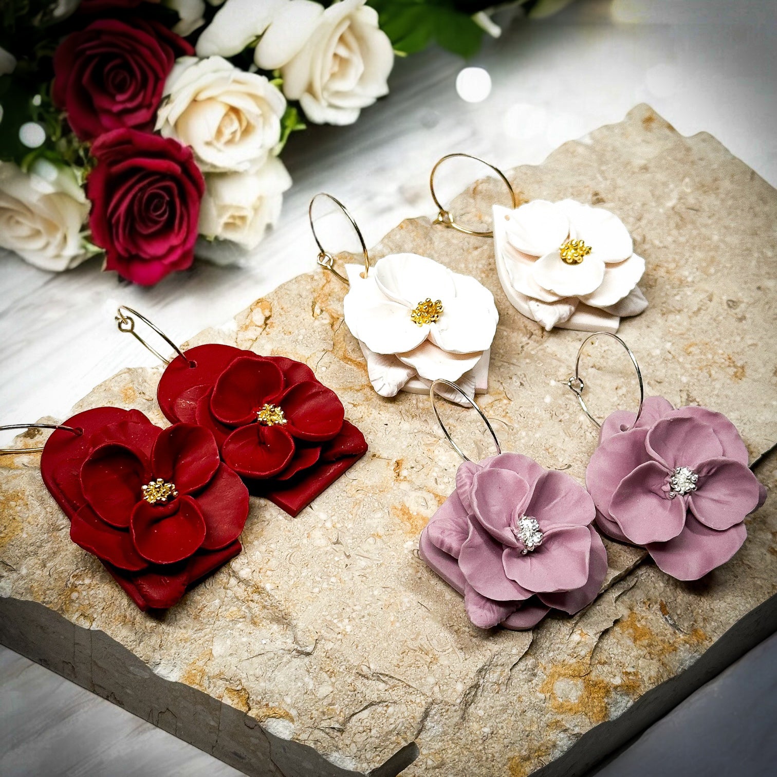 The Dappled Wood Hearts & Crafts 2025 class sample earrings displayed on textured stone. Includes red, cream, and violet floral polymer clay earrings with gold and silver details. A bouquet of roses in the background adds a romantic Galentines touch.