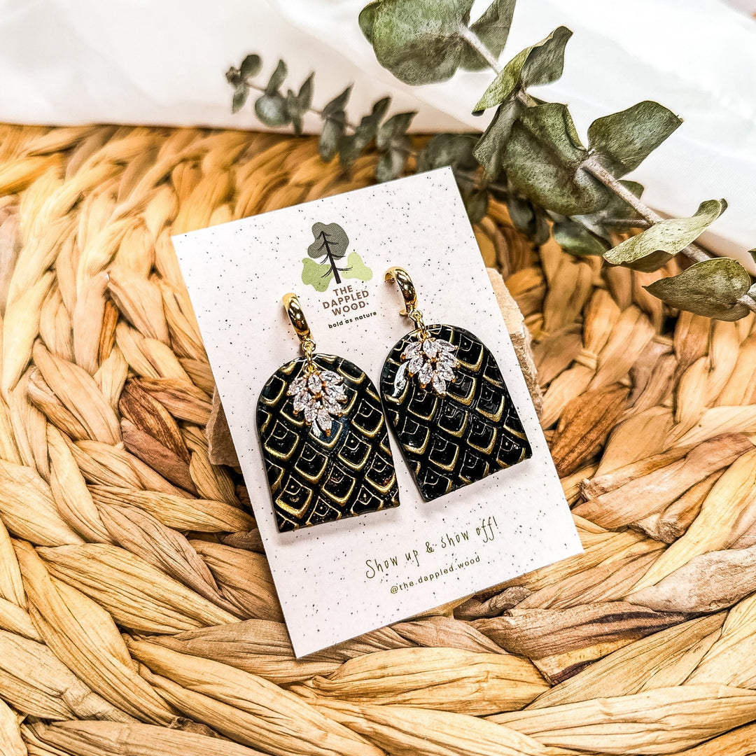 Closeup photo of the Hermione brass scale earrings from The Dappled Wood. Black and gold scale patterned arches with textured leaf details displayed on a branded earring card.