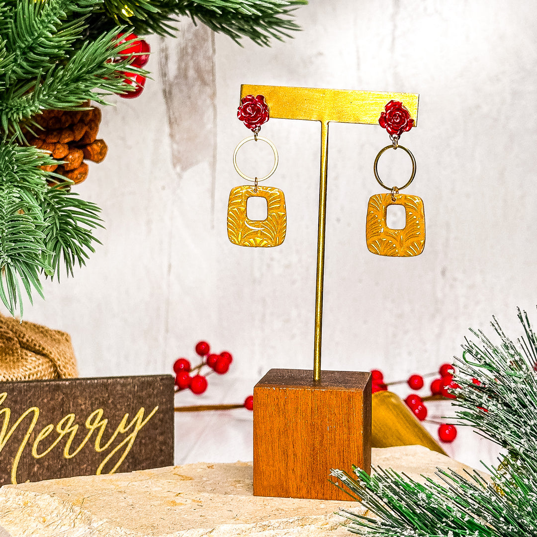 The Dappled Wood Belle earrings displayed on a stand, featuring red floral details and geometric gold dangles. Elegant, lightweight statement jewelry for weddings and holiday celebrations.