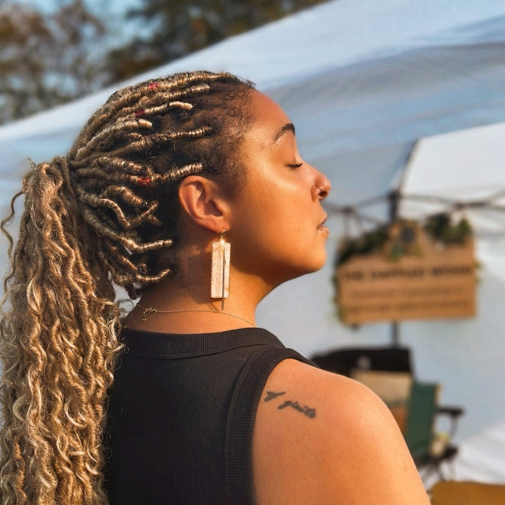 Customer wearing the Ivory Coast rectangle earrings by The Dappled Wood, featuring lightweight gold accented statement drops styled outdoors at a market booth in warm sunlight.
