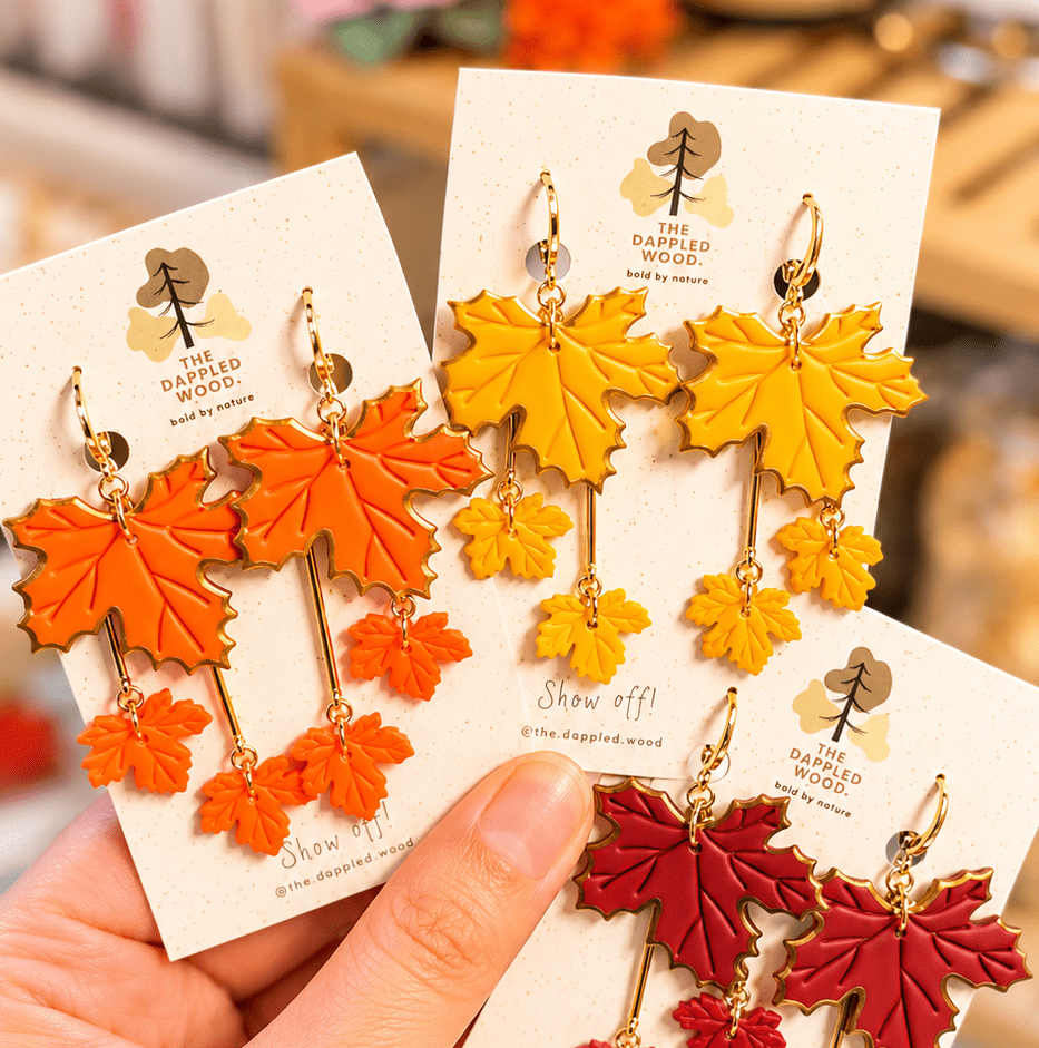 Laurel tiered maple leaf earrings in orange, yellow and red displayed on cards held in a hand.