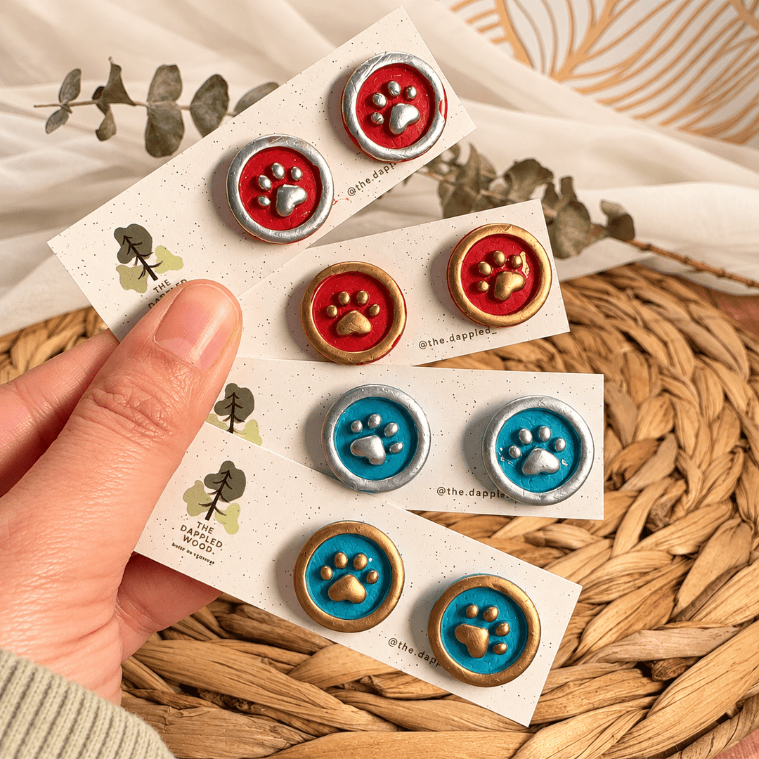 Maisie paw-print stamp stud earrings in red and teal with gold or silver details on cards.