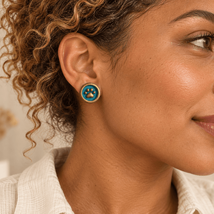 AI-generated styling preview of Maisie teal and gold paw-print stud earrings on a model with honey curls; scale illustrative.