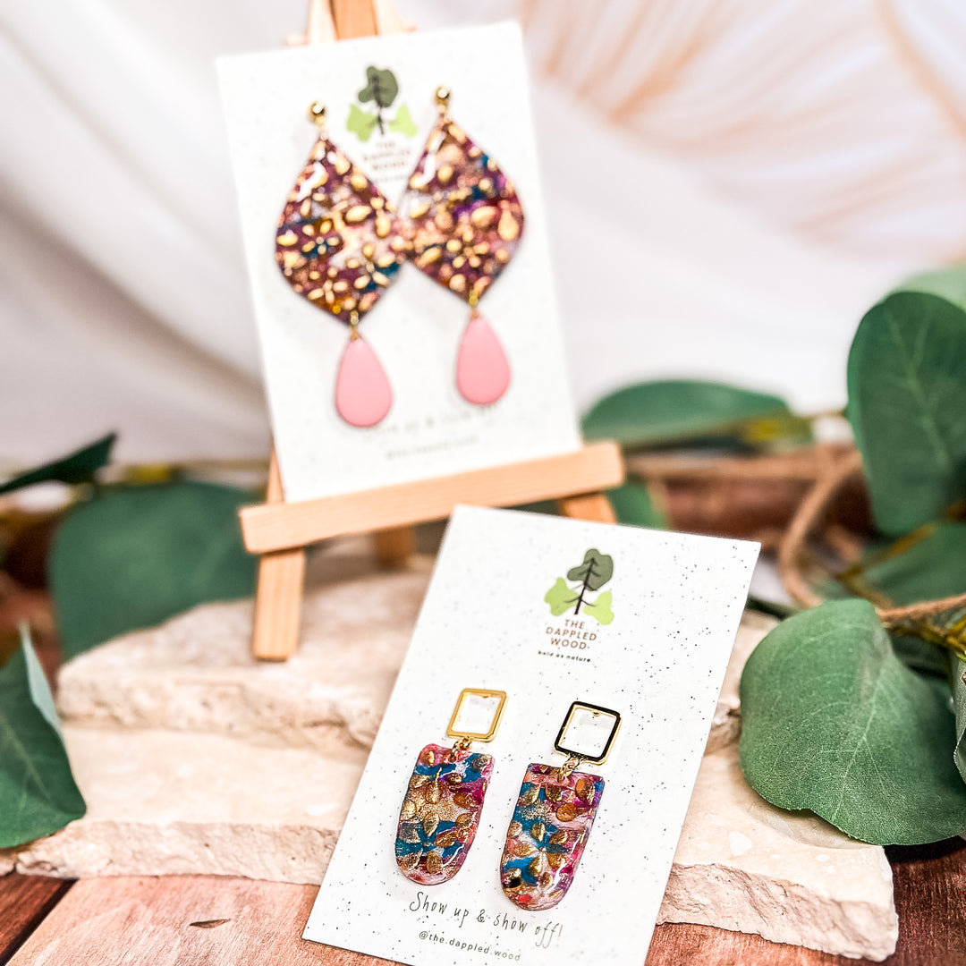 The Dappled Wood Dorothy earrings collection, showcasing floral designs in teardrop and rectangle styles with shimmering gold accents. A bold yet elegant accessory for New Year's parties.