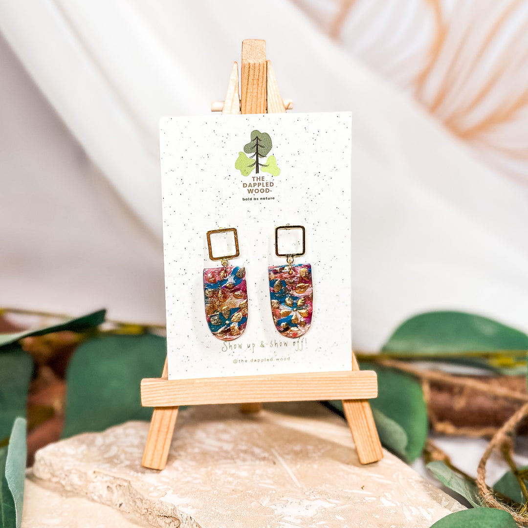 The Dappled Wood Dorothy floral rectangle earrings featuring gold shimmer accents, displayed on a festive backdrop. Elegant statement jewelry for ringing in the new year.