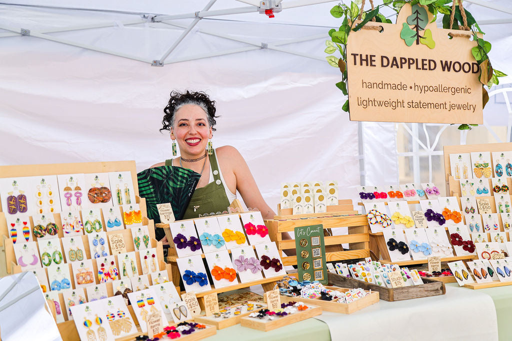 Brittany Arnett, owner of The Dappled Wood, standing at her market booth with displays of handmade lightweight statement jewelry arranged in bright colorful collections.