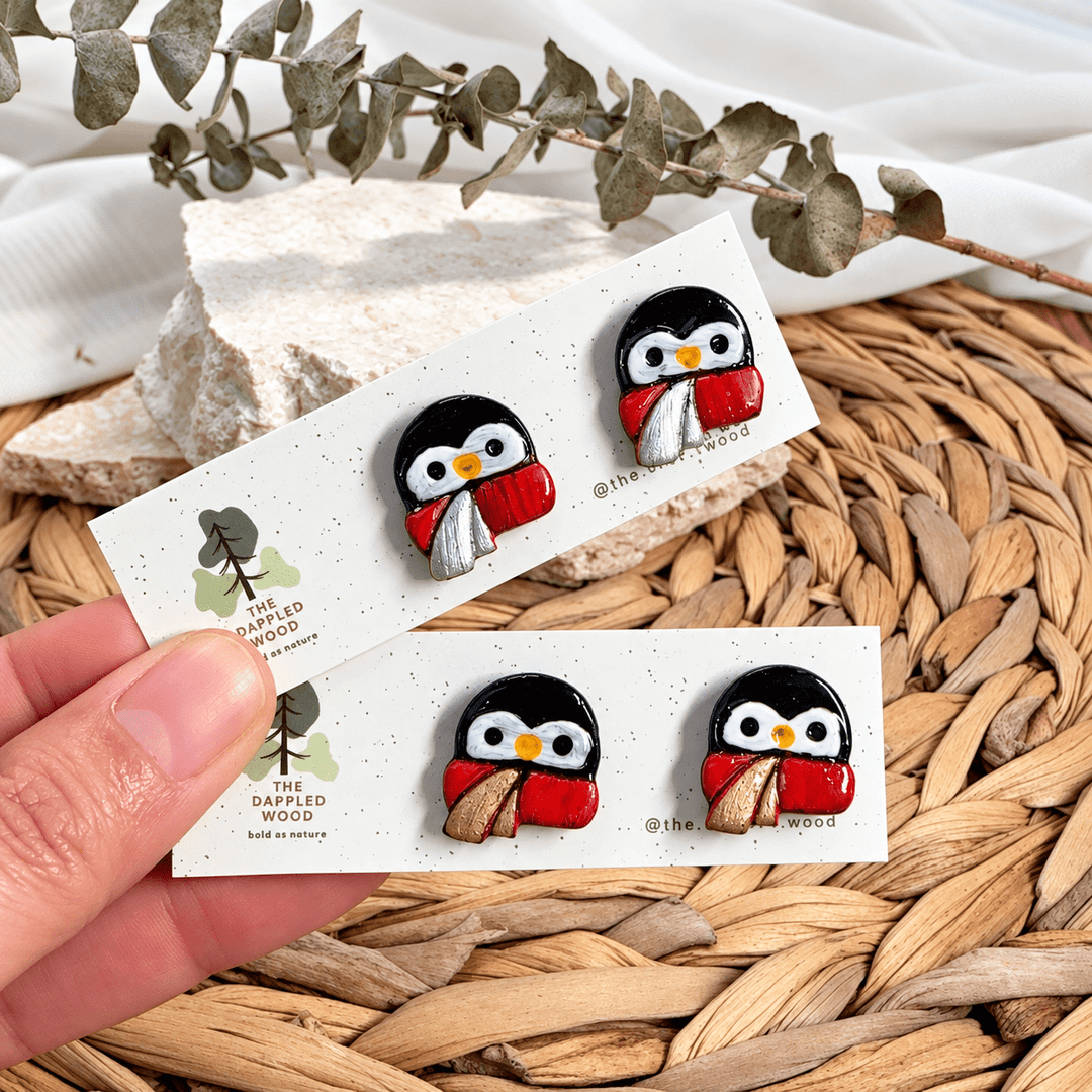Pippa hand-painted penguin stud earrings with red scarves and gold or silver accents on display cards.