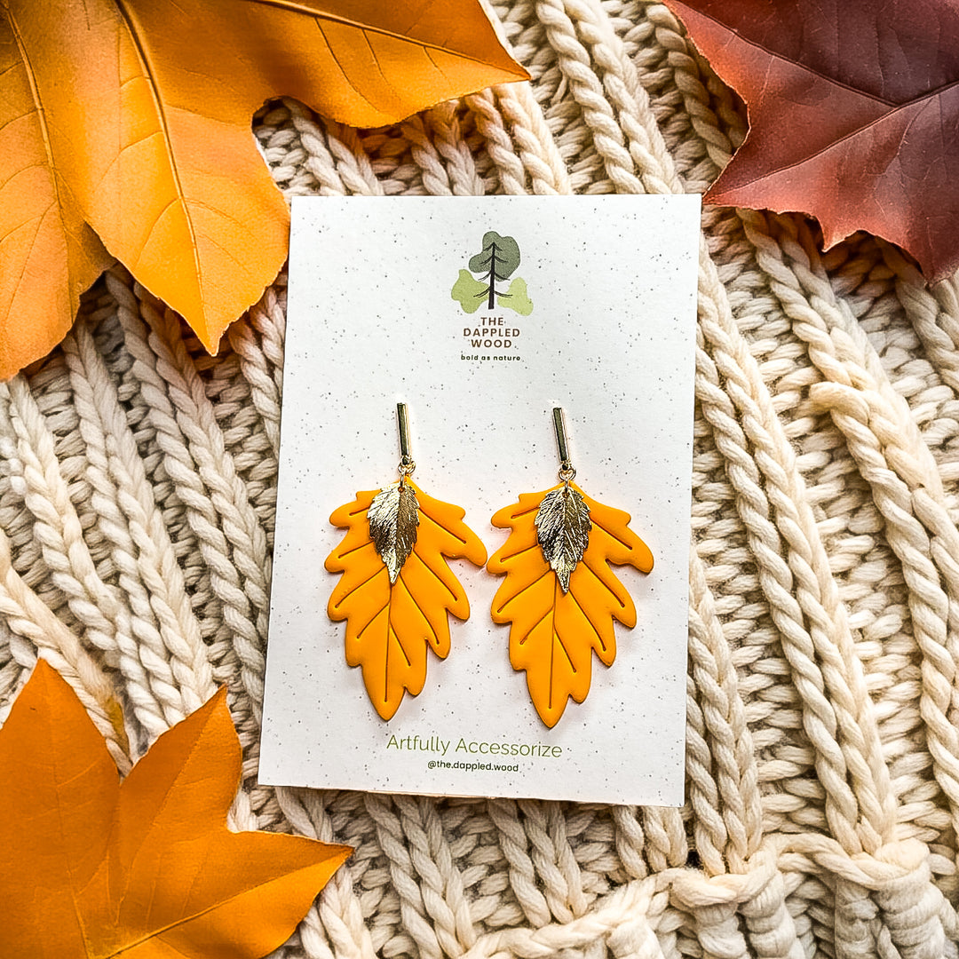 Solana earrings in Sunflower Yellow by The Dappled Wood displayed on a branded card, featuring bright yellow layered leaves with gold accents on a cream knit background with fall foliage.