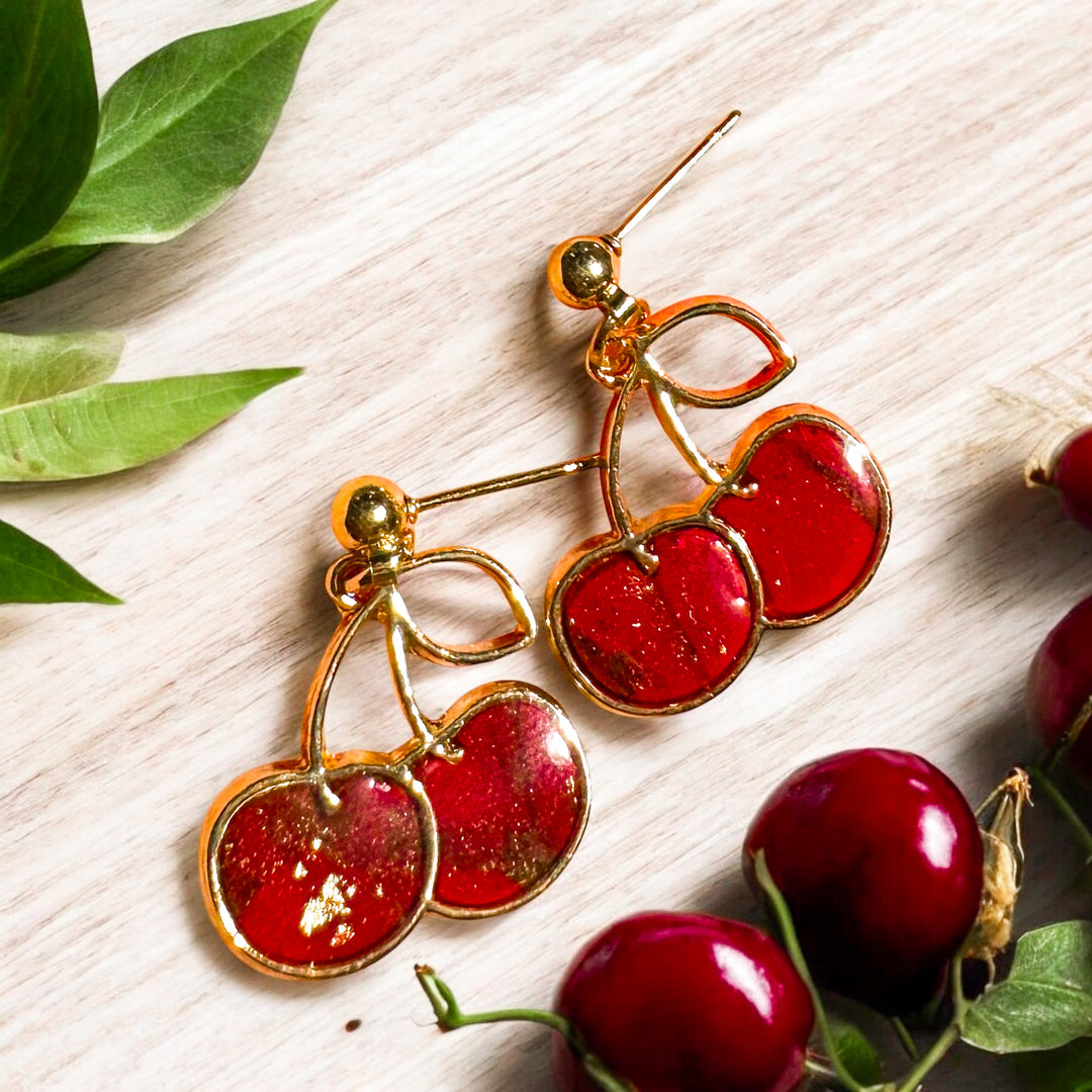 A close-up of gold-framed cherry earrings handmade with vibrant red clay, and accented with delicate gold leaf detailing. Styled on a light wooden surface with fresh cherries and green leaves. Part of The Dappled Wood's Spring 2025 earring collection.