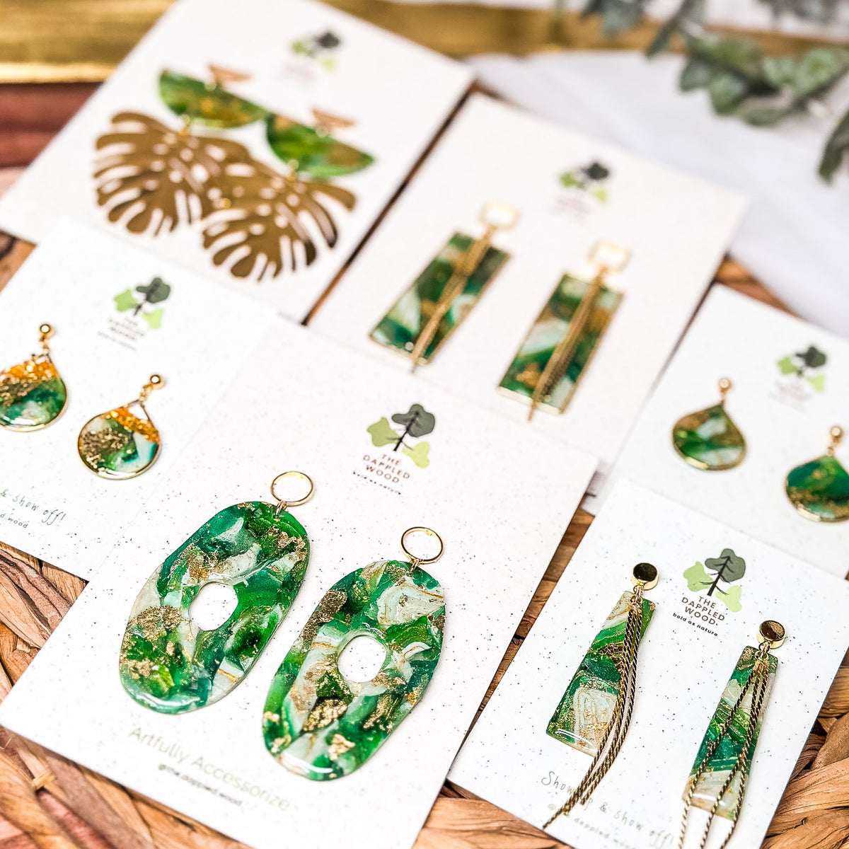 The Dappled Wood Gilded Grove Collection flat lay featuring a selection of handmade green and gold statement earrings, including the Chloe earrings, displayed on branded earring cards, showcasing nature-inspired lightweight hypoallergenic jewelry for Spring 2025.