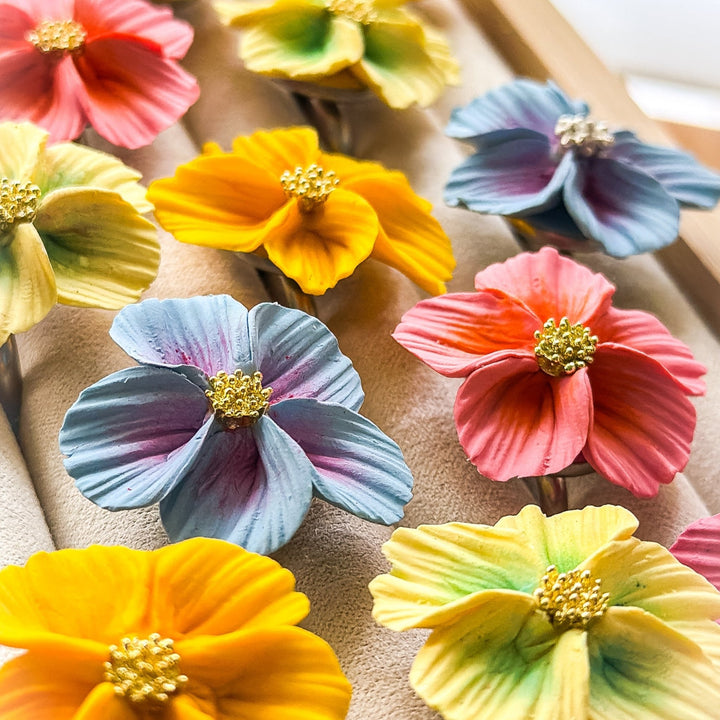 The Dappled Wood Isadora floral cocktail rings, featuring vibrant flower petals in shades of blue, pink, and yellow with delicate gold centers, perfect as statement jewelry for spring and summer accessorizing.
