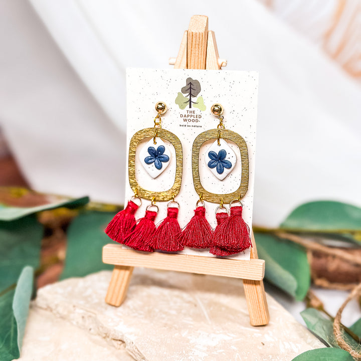 The Dappled Wood Bobbi earrings, featuring deep blue floral accents on an ivory base framed by textured gold ovals. Finished with bold red tassels for a patriotic summer statement.