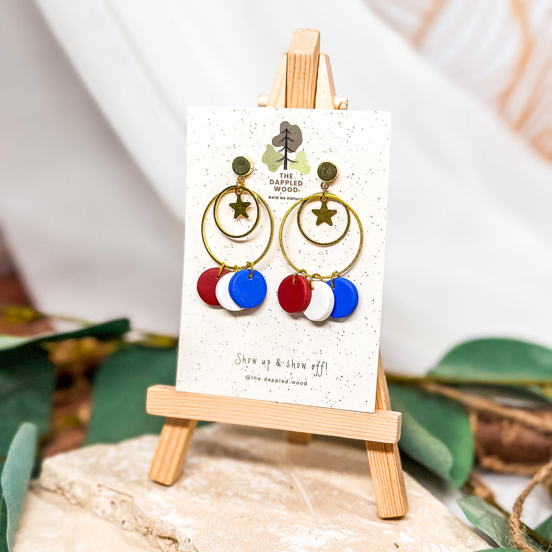 The Dappled Wood Petunia earrings, featuring antique gold hoops with star charms and red, white, and blue polymer clay dangles. A festive and lightweight statement for patriotic celebrations.