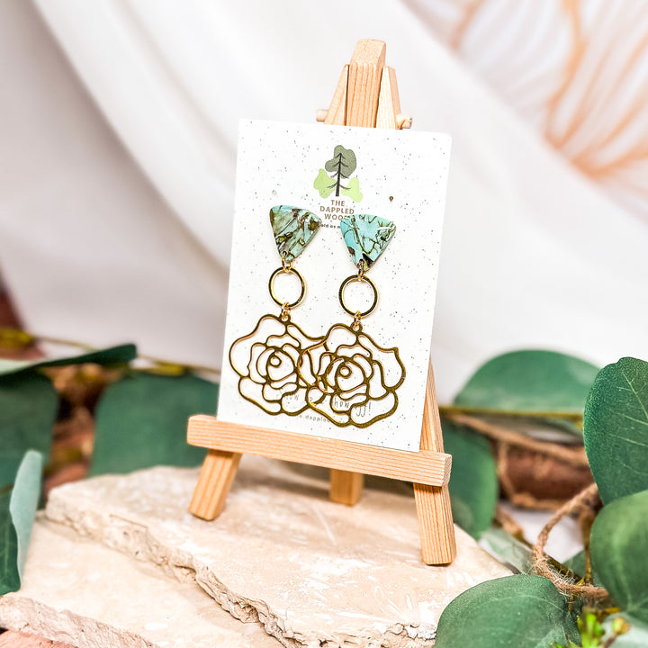  The Dappled Wood Dusty Turquoise earrings, featuring handcrafted faux stone polymer clay tops in a marbled turquoise pattern with brass rose charms. Set on 18k gold-filled posts for a lightweight yet elegant statement piece.