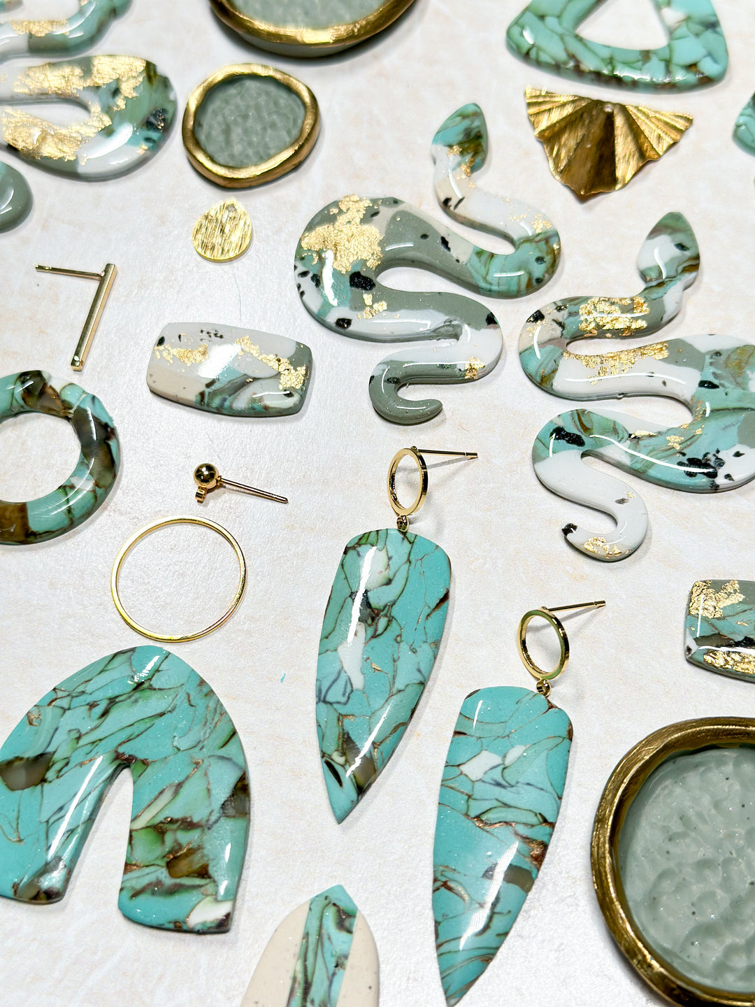 The Dappled Wood's Dusty Turquoise Jewelry Collection flat lay. A curated selection of earrings in marbled blue-green hues with gold accents, featuring various shapes, textures, and premium hypoallergenic metals.