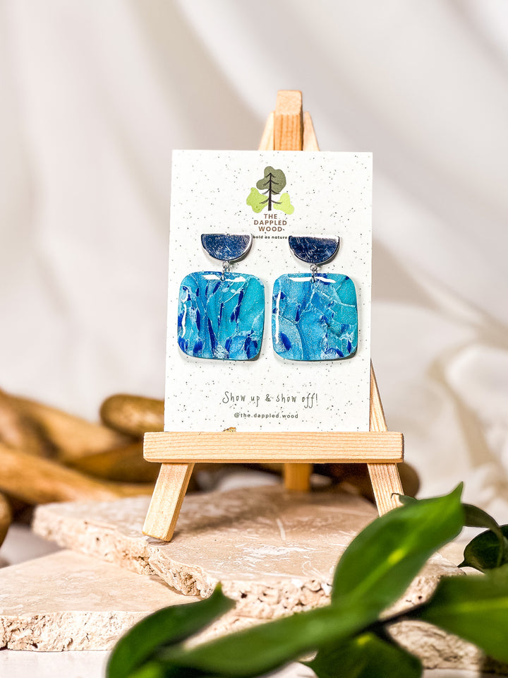 Earrings light blue in color, featuring a square design with a silver half-moon post. A modern take on faux larimar earrings with a sleek and minimalistic style. Part of The Dappled Wood's Poolside collection from summer 2024.