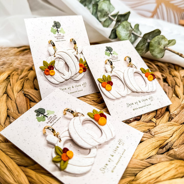Flatlay photo of the Sylvia pumpkin earrings from The Dappled Wood displayed on branded earring cards. White textured pumpkin hoops with leaf and berry details arranged on a woven surface.