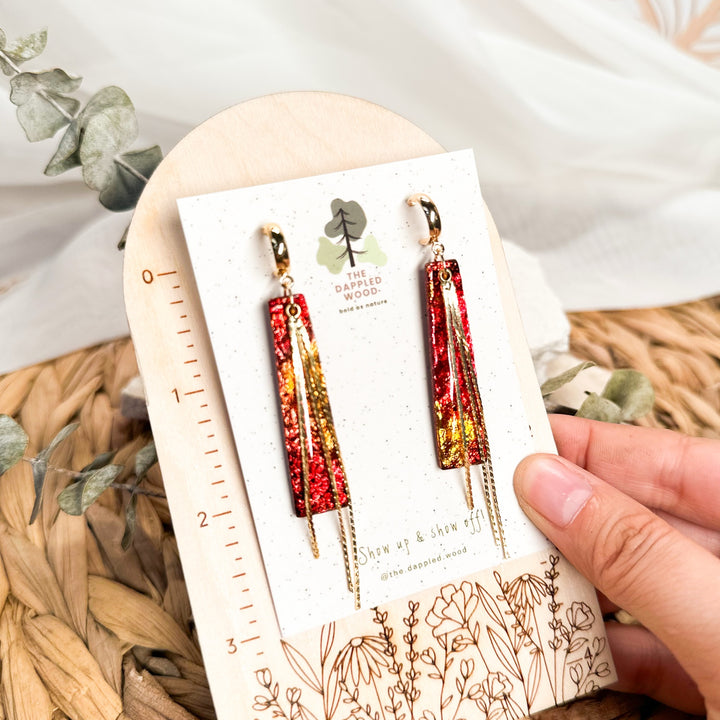 Ximena red and gold faux fire opal tassel earrings on a card beside an inch ruler.