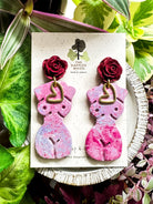 Marbled lavender and pink polymer clay earrings in the shape of a natural, curvy woman with a gold heart pendant, hanging from deep red rose posts. Earrings are showcased on a branded 'The Dappled Wood' card surrounded by lush greenery.