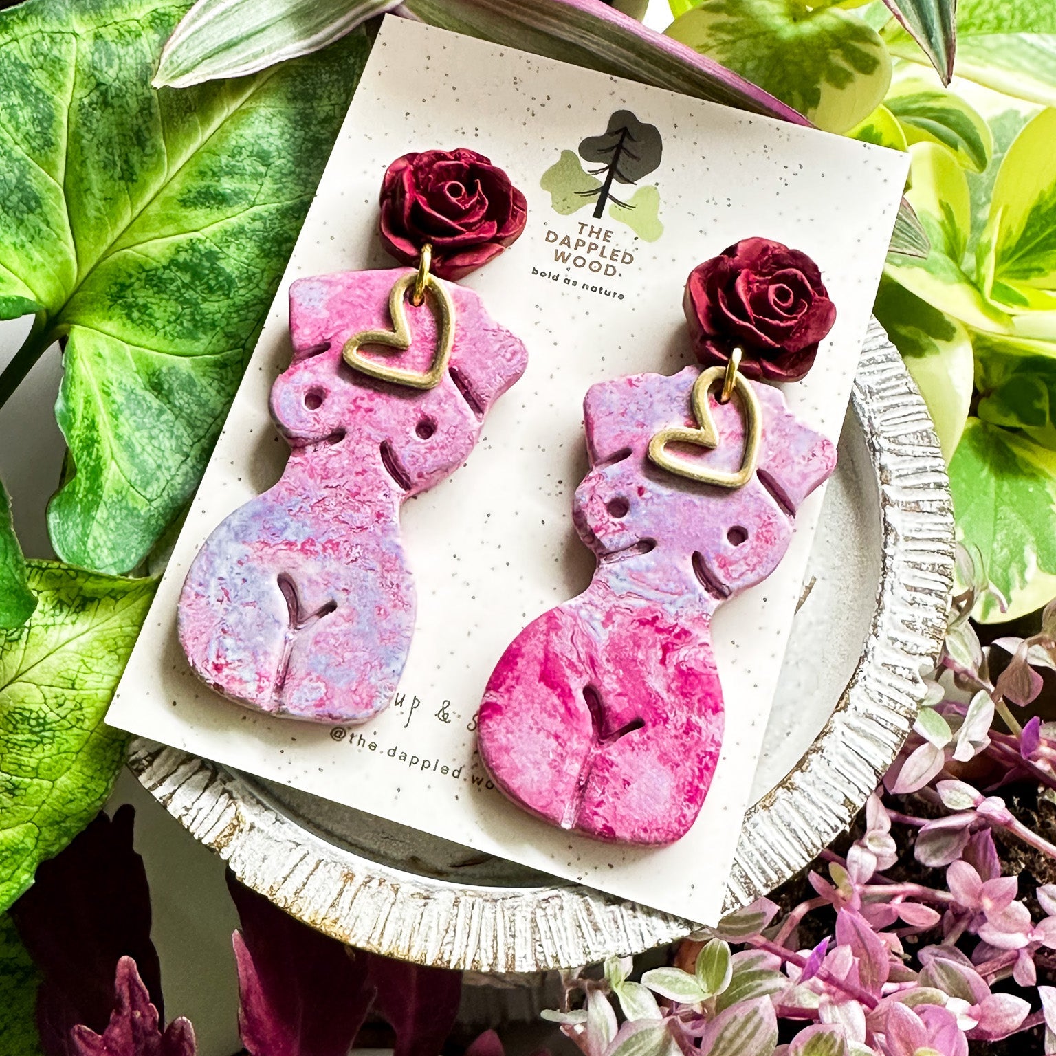 Marbled lavender and pink polymer clay earrings in the shape of a natural, curvy woman with a gold heart pendant, hanging from deep red rose posts. Earrings are showcased on a branded 'The Dappled Wood' card surrounded by lush greenery.