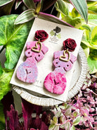 Marbled lavender and pink polymer clay earrings in the shape of a natural, curvy woman with a gold heart pendant, hanging from deep red rose posts. Earrings are showcased on a branded 'The Dappled Wood' card surrounded by lush greenery.