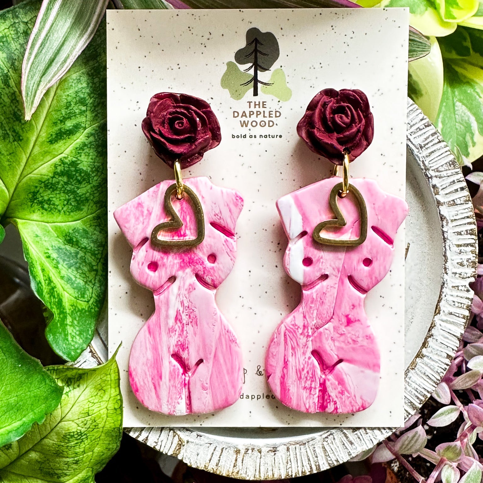 Marbled white and pink polymer clay earrings in the shape of a natural, curvy woman with a gold heart pendant, hanging from deep red rose posts. Earrings are showcased on a branded 'The Dappled Wood' card surrounded by lush greenery.